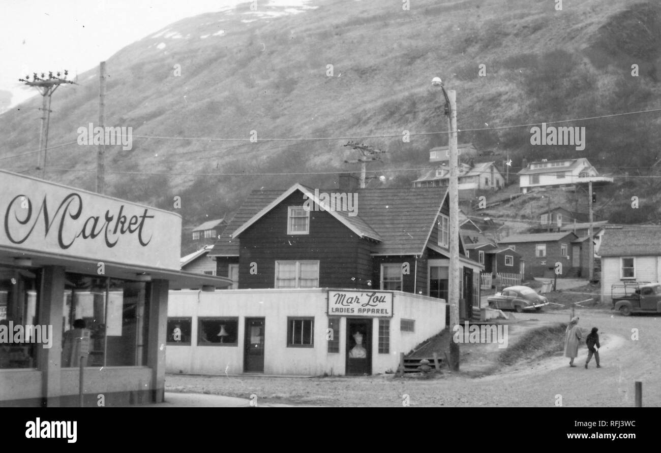 Black and white photograph of commercial and domestic buildings in a small town in Alaska; with a concrete, deco storefront at left with the sign 'Market, ' and a low white building in the foreground with the sign 'Mar' Lou Ladies Apparel'; a warmly dressed boy and a woman wearing a coat and scarf, walk up a dirt road at right, and several wooden buildings and snow-covered slopes are visible in the background; photographed during a hunting and fishing trip located in Alaska, 1955. () Stock Photo