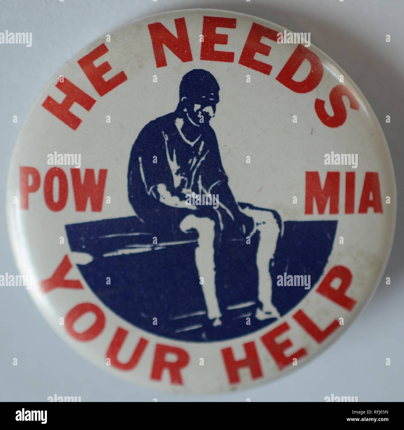 Red, white and blue pinback protest button with silhouette of a prisoner of war sitting forlornly in a cell, with text reading POW MIA He Needs Our Help, advocating for return of Prisoners of War and those Missing in Action during the Vietnam War, 1970. () Stock Photo