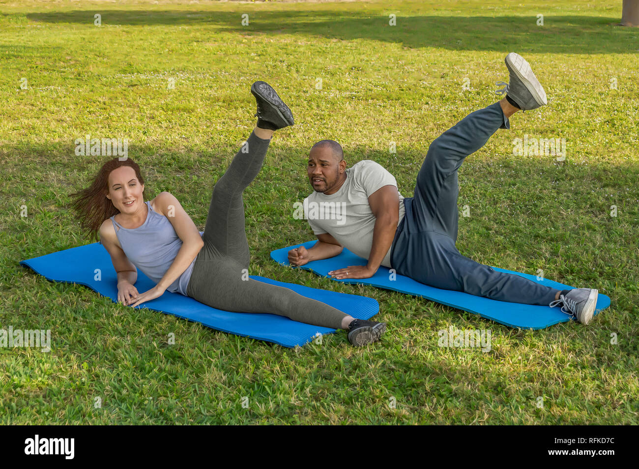 An interracial couple stretches together in the morning at the downtown park. They start by doing leg lifts on blue yoga mats. Stock Photo