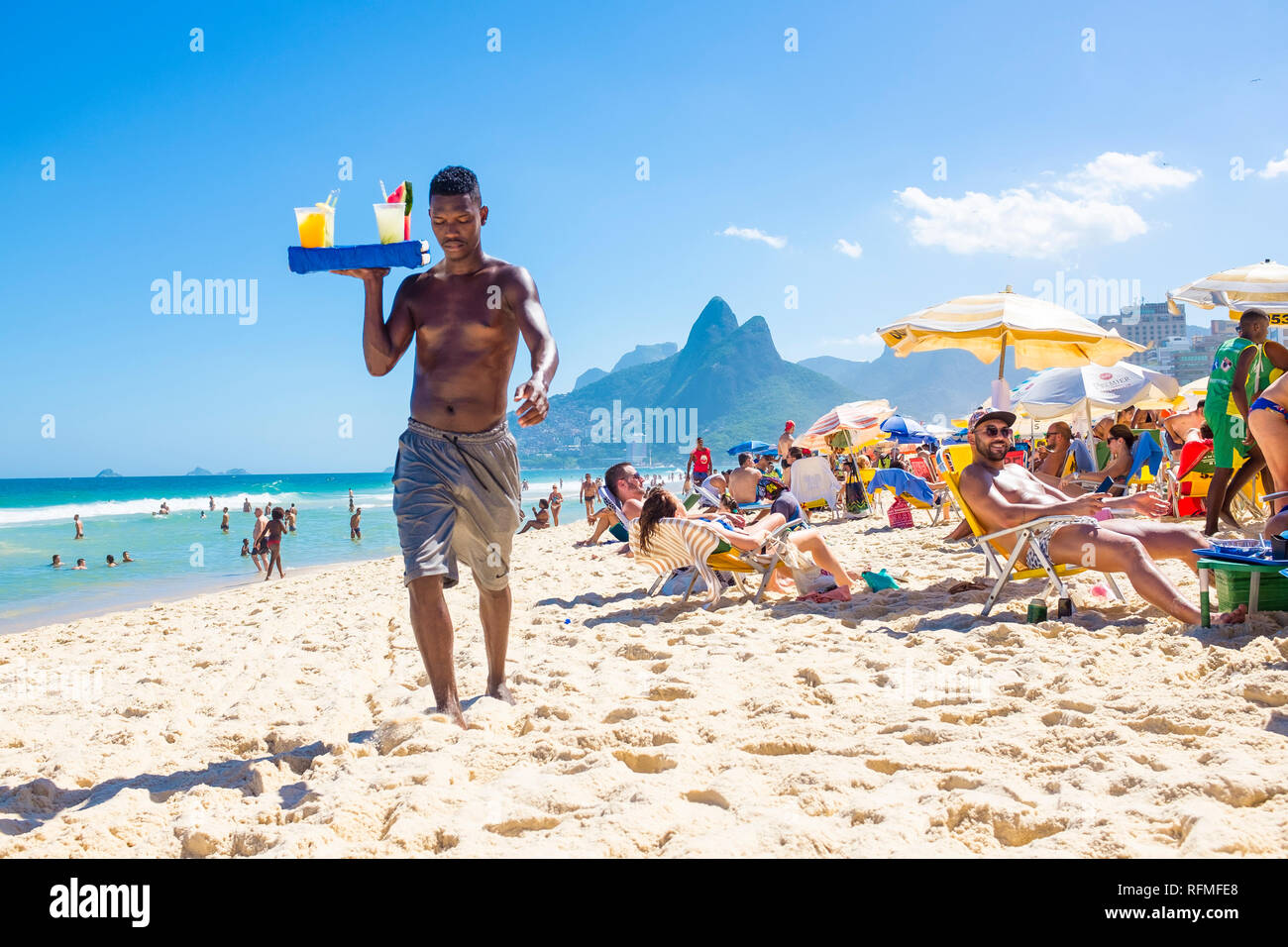 RIO DE JANEIRO - FEBRUARY, 2018: An unlicensed Brazilian beach vendor carries a tray of homemade caipirinha cocktails past beachgoers on Ipanema Beach Stock Photo
