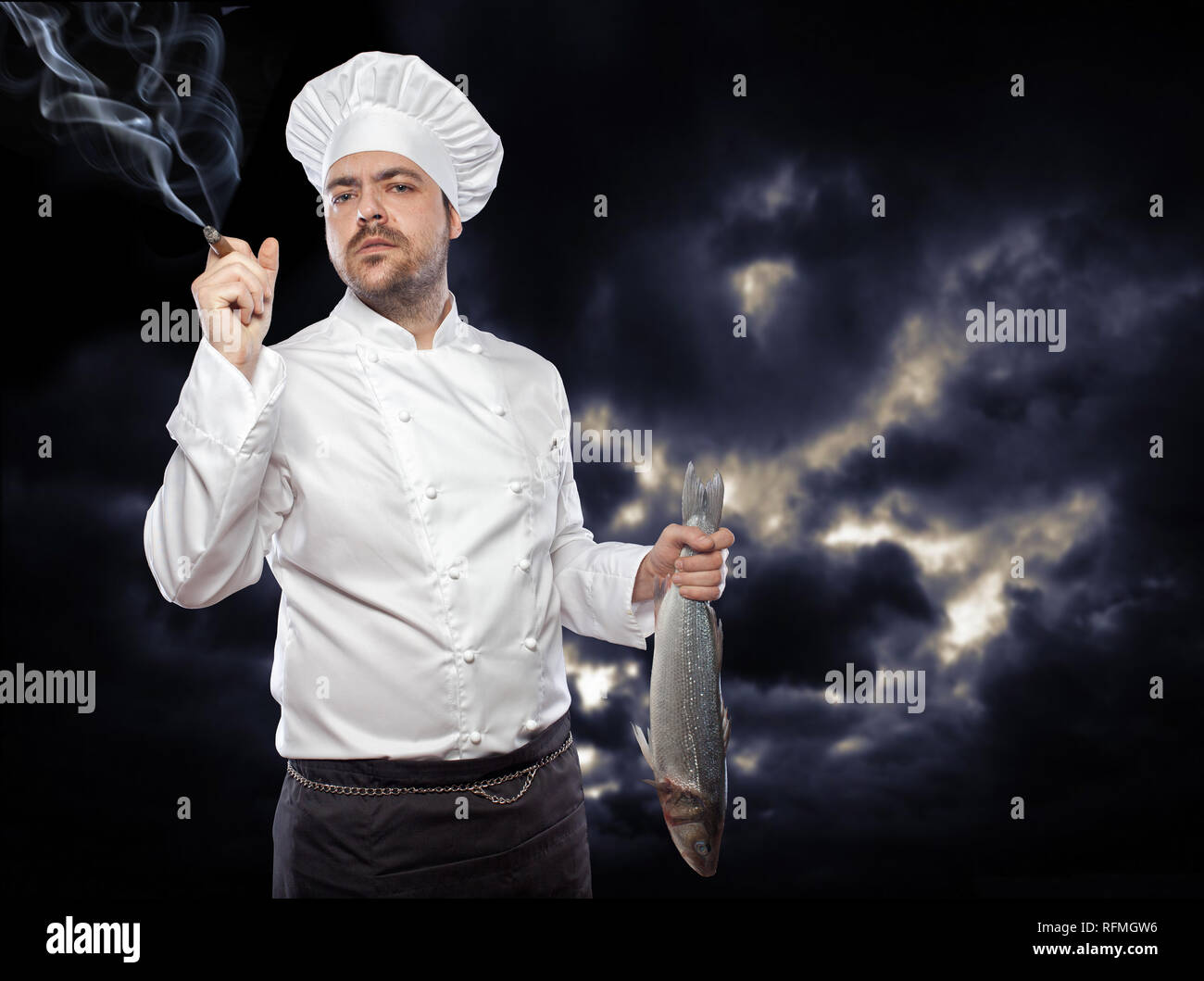 Young chef with sea bass fish smoking cigar against dark sky Stock