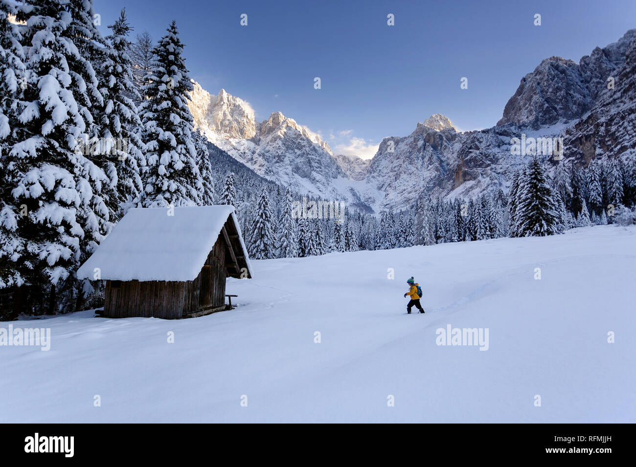 Winter outdoors can be fairytale-maker for children, young boy hiking in snow to a wooden chalet, mountains in light in the background Julian Alps Stock Photo