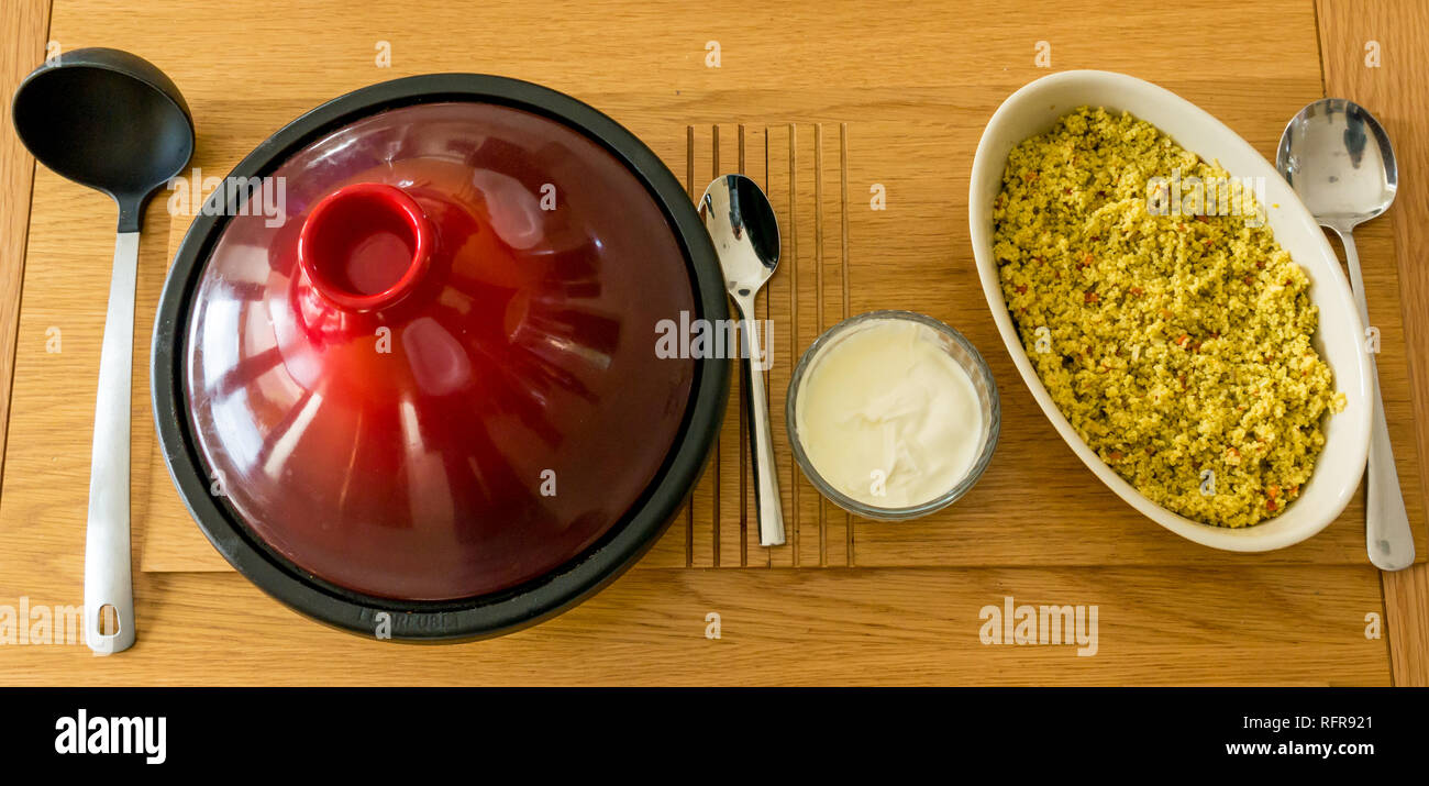 Red ceramic and cast iron tagine on oak table with serving ladle, couscous, yoghurt and white crockery Stock Photo