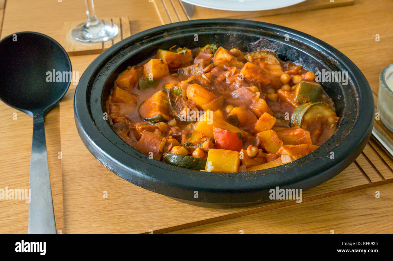 Vegetarian meal of vegetable tagine in cast iron pot with butternut squash, courgette, onions, peppers and serving ladle on oak table Stock Photo