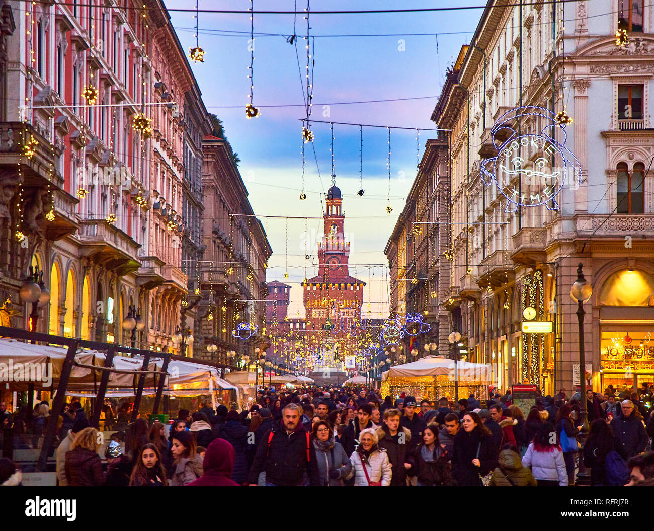 People walking on Via Dante street illuminated by christmas lights at nightfall with the Filarete Tower of the Castello Sforzesco in background. Milan Stock Photo