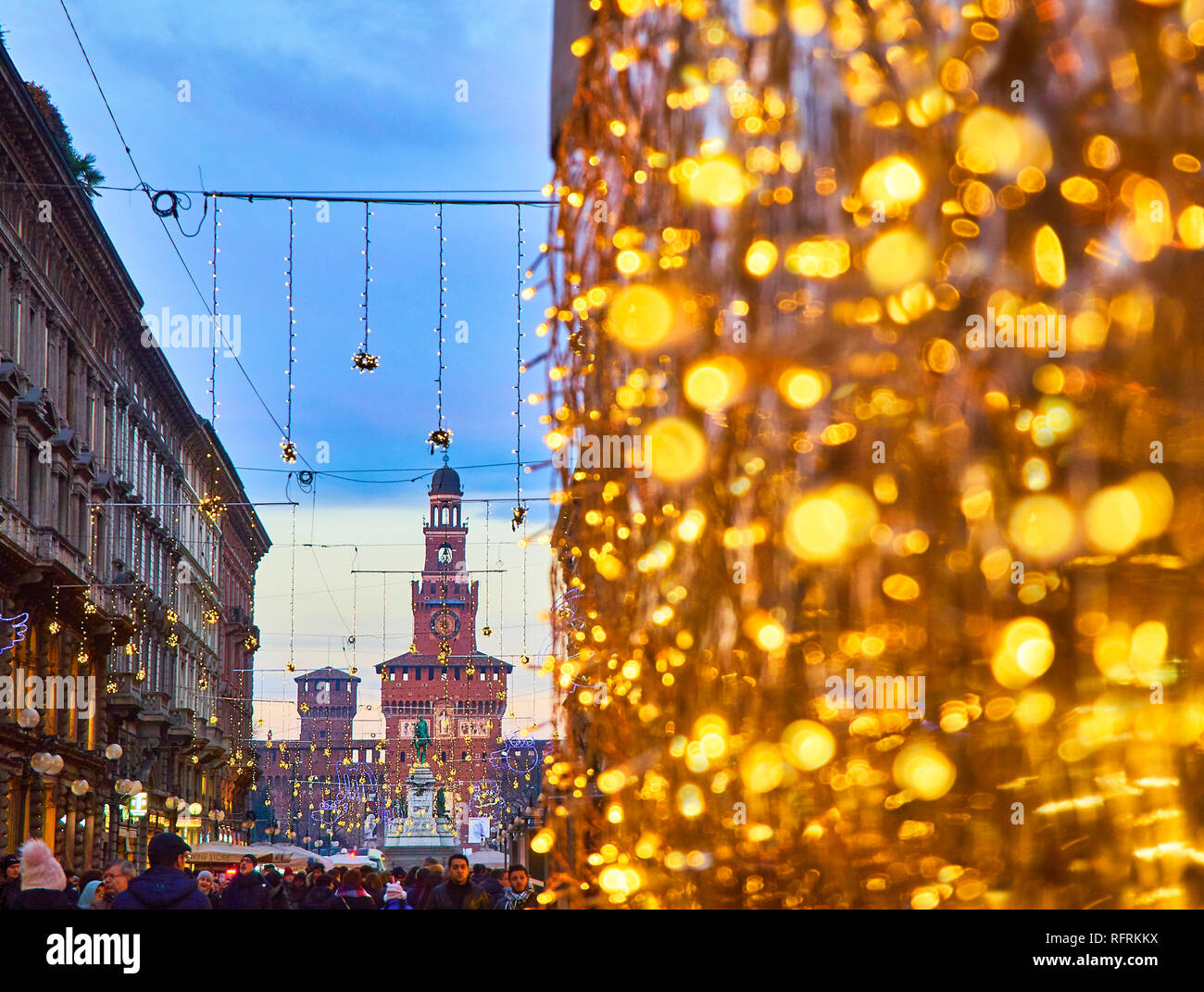 People walking on Via Dante street illuminated by christmas lights at nightfall with the Filarete Tower of the Castello Sforzesco in background. Milan Stock Photo