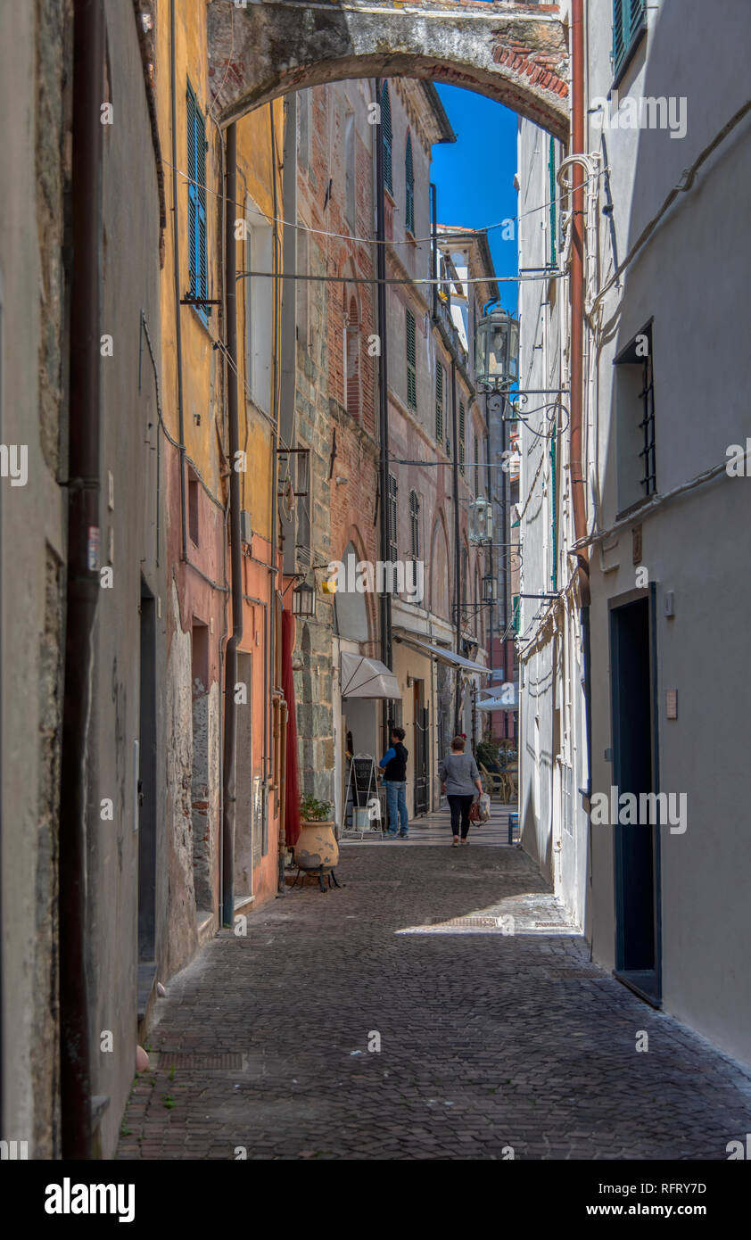 Narrow cobbled lane between historic low-rise buildings in Noli, Liguria , Italy with local pedestrians in the distance going about everyday life Stock Photo