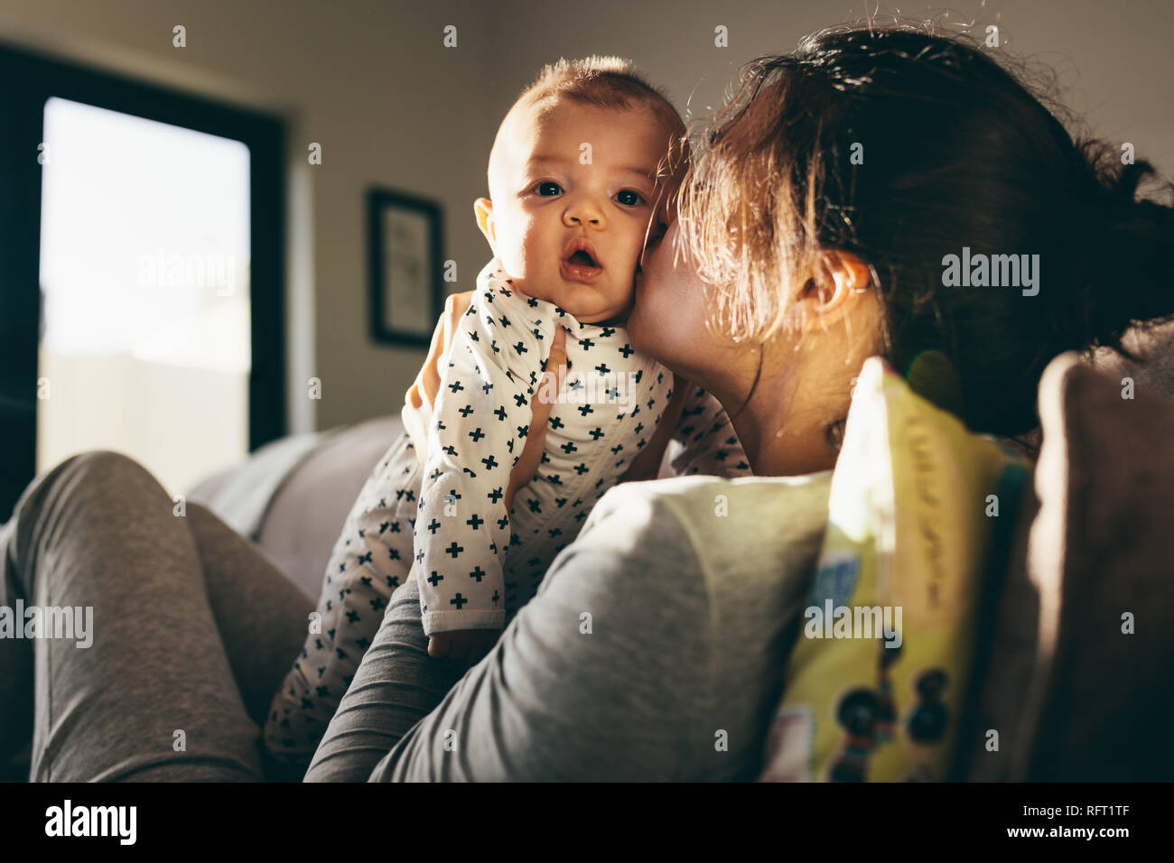 Young mother kissing her baby sitting on a couch at home. Close up of a woman holding her infant kid and pampering him. Stock Photo