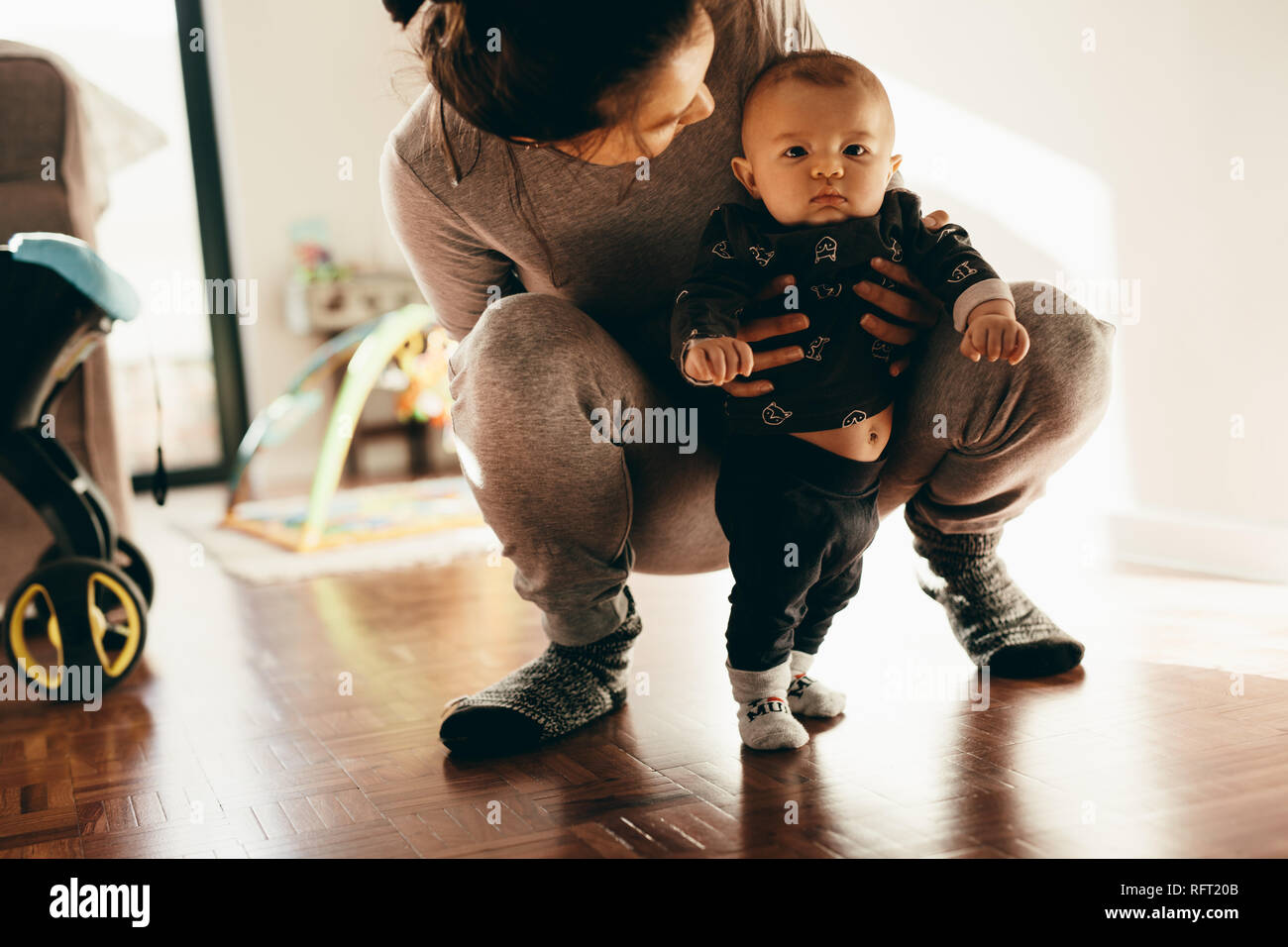 Close up of a woman playing with her baby squatting on the floor and holding him. Mother making her baby stand on floor at home. Stock Photo