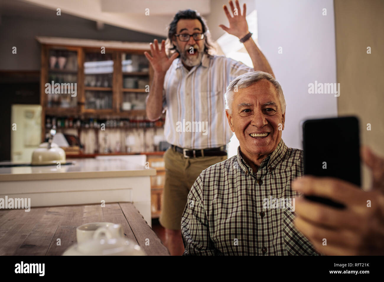 Happy senior man taking selfie with friend standing at back with his hands raised. Two elderly men enjoying taking selfie with mobile phone at home. Stock Photo