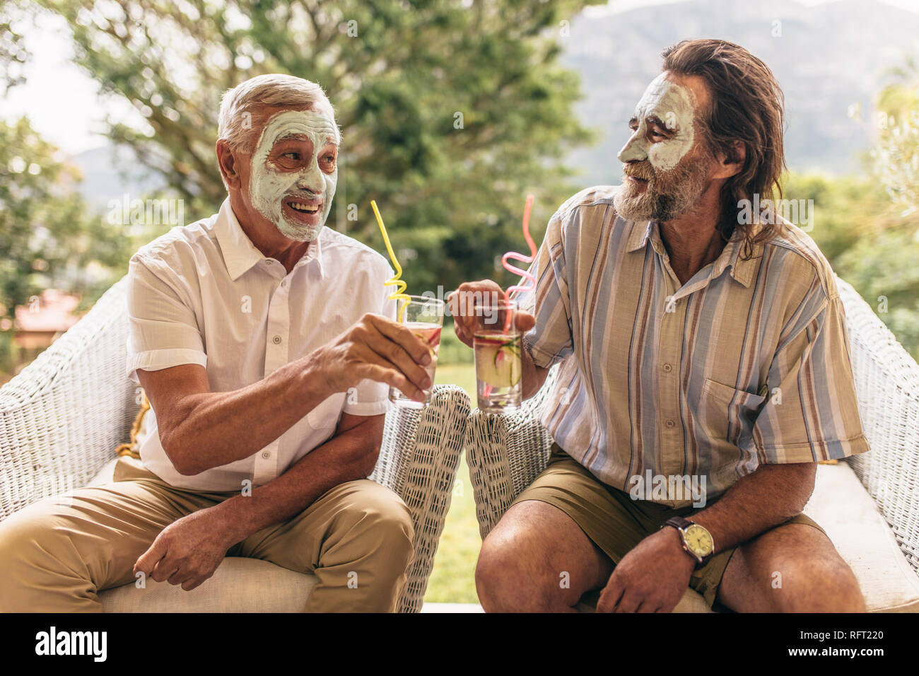 Two elderly friends with facial clay mask on clinking juice glasses. Senior men sitting on chair with spa face mask having juice. Stock Photo