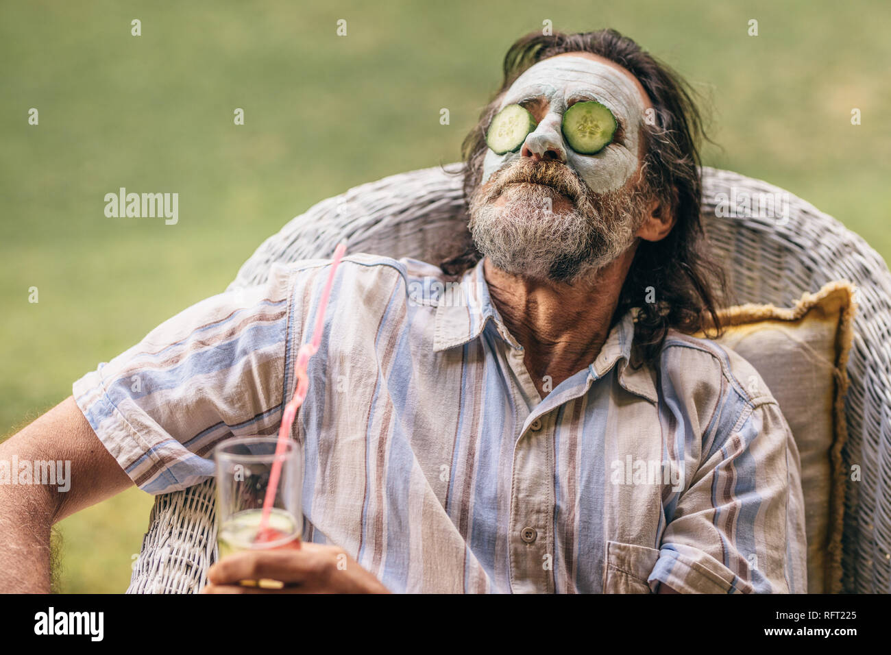 Bearded elderly man sitting on chair with clay mask and cucumber slice on face. Retired senior man taking care of his skin at old age. Stock Photo