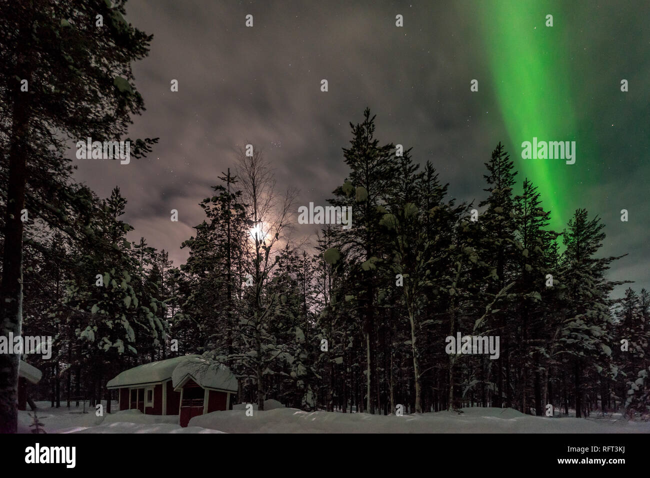 The northern lights and the moon illuminates the snow covered forest and a log cabin in Finnish Lapland, Pyha, Finalnd. Stock Photo