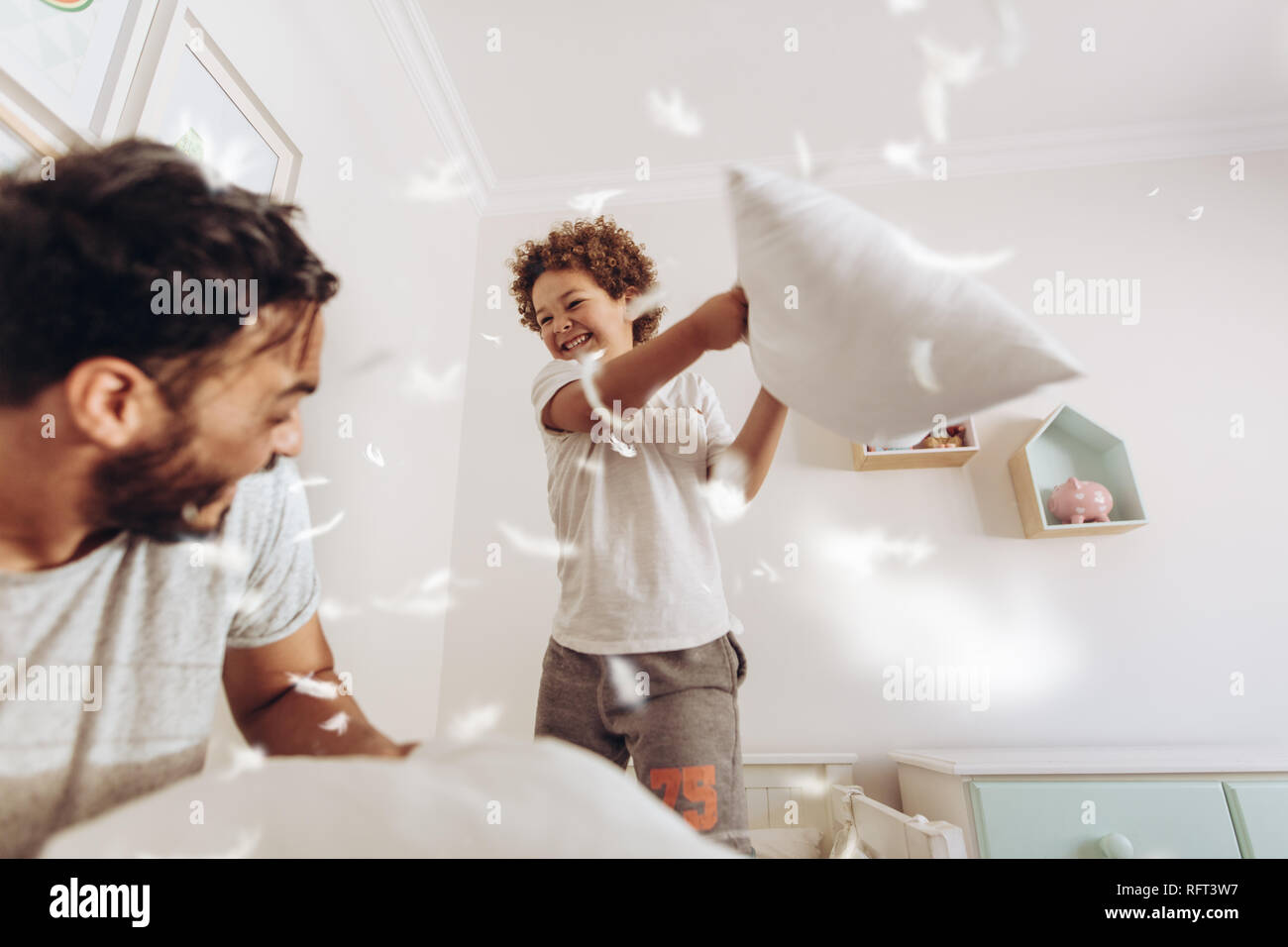 Happy father and son having a pillow fight on bed with feathers flying around. Father and son having fun playing at home. Stock Photo