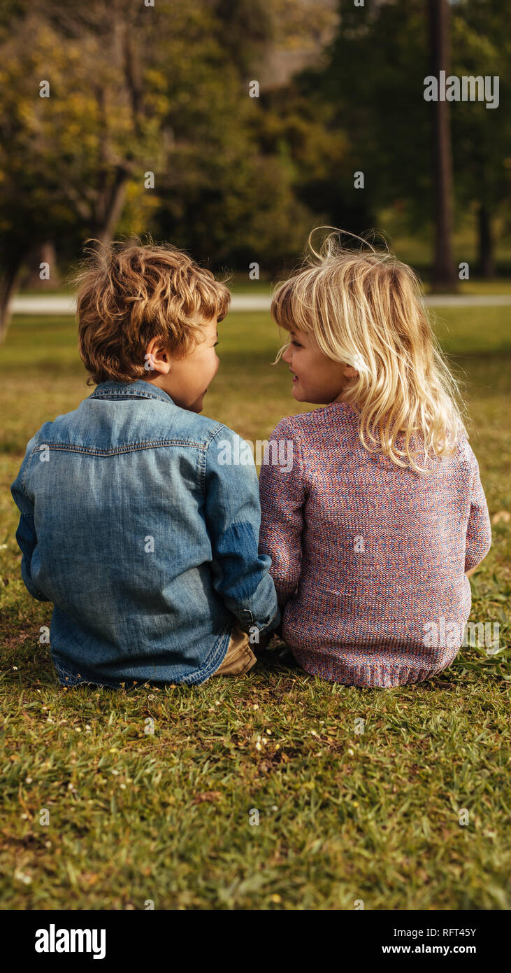 Rear shot of siblings sitting on the grass and looking at each other faces. Little sister sitting with her brother at park. Stock Photo