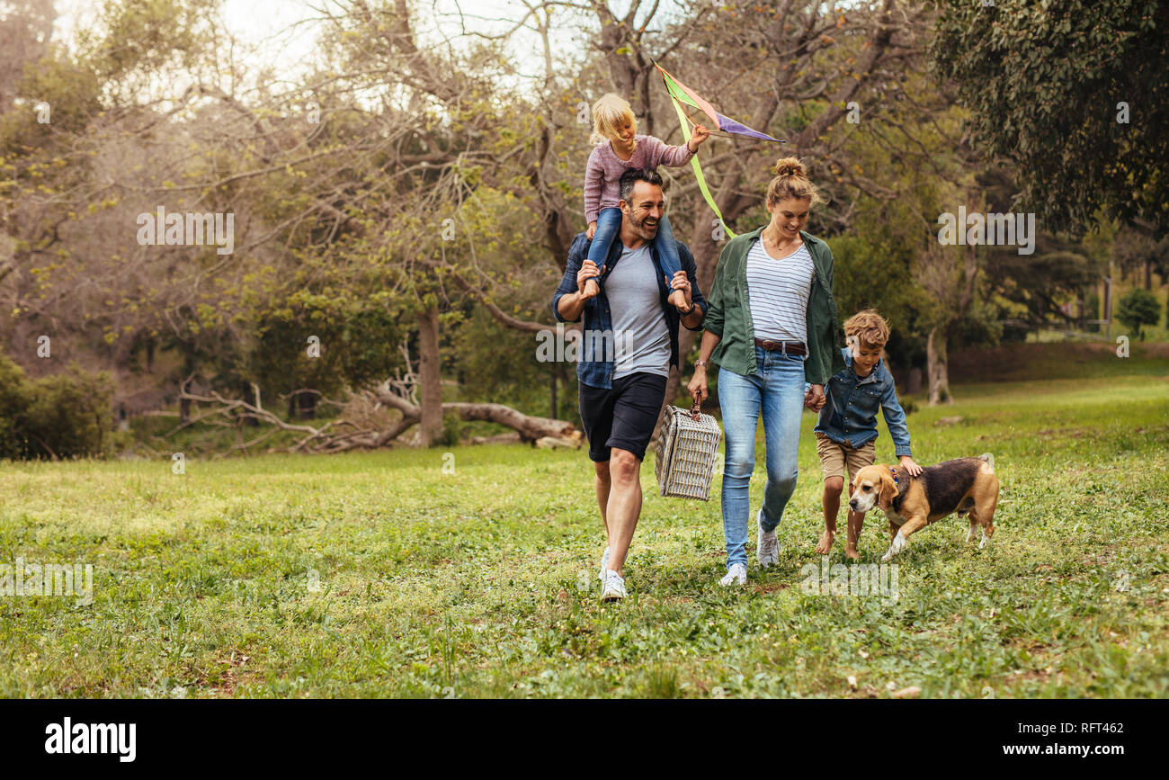 Happy couple with their two children and dog walking in park. Man carrying little girl with kite on shoulders and woman carrying a picnic basket with Stock Photo