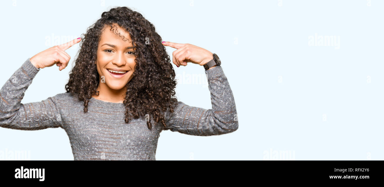 Young beautiful woman with curly hair wearing grey sweater Smiling pointing to head with both hands finger, great idea or thought, good memory Stock Photo