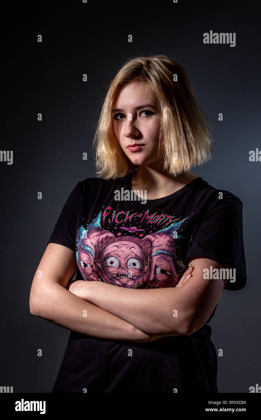 Photo of a serious young American blonde girl with arms crossed in a tattoo Studio with a pulsating light against a gray wall, a look into the camera Stock Photo