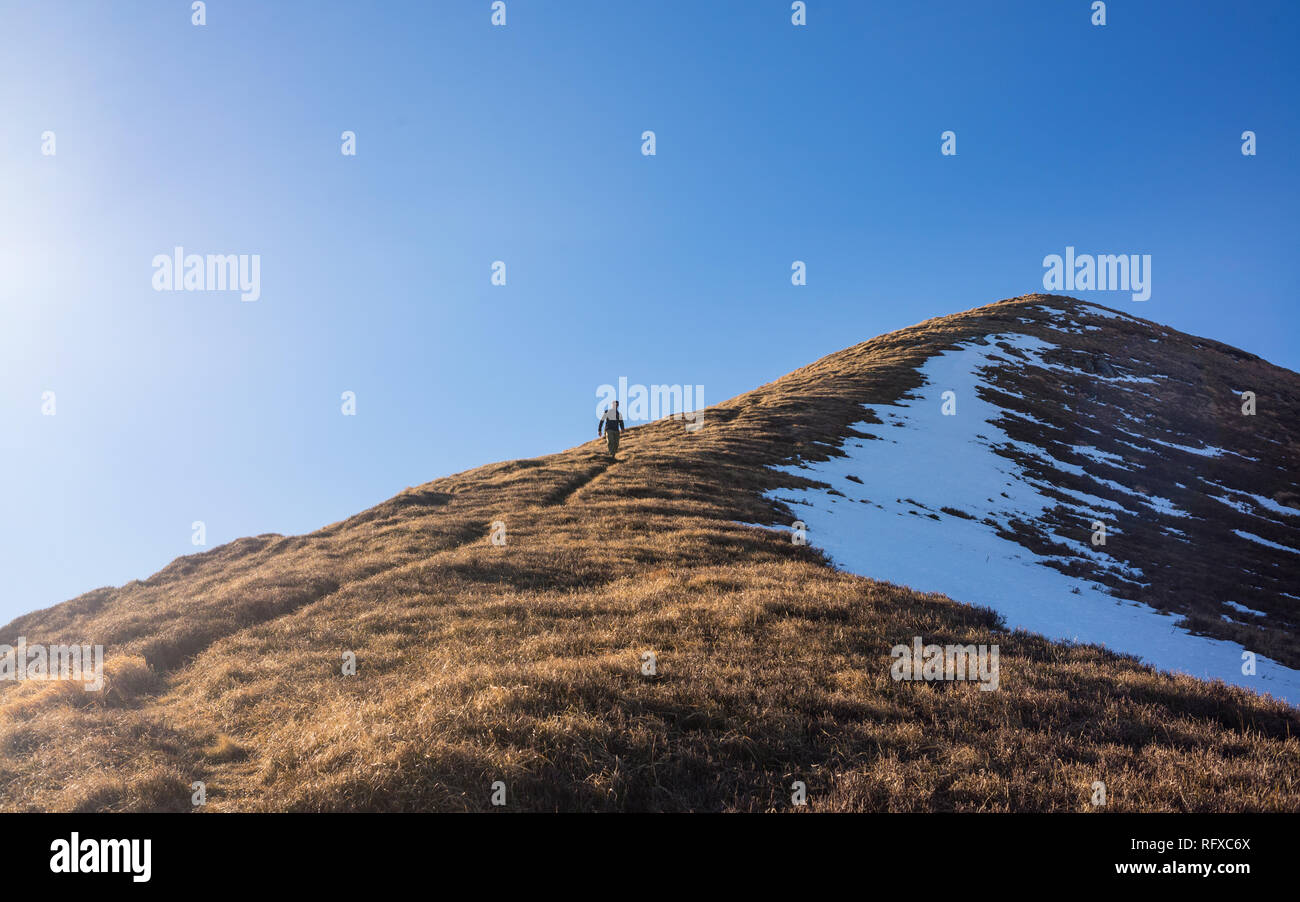 One Man hiker hiking alone climbing down a crest ridge of steep mountain path in winter. Taken from distance. Stock Photo