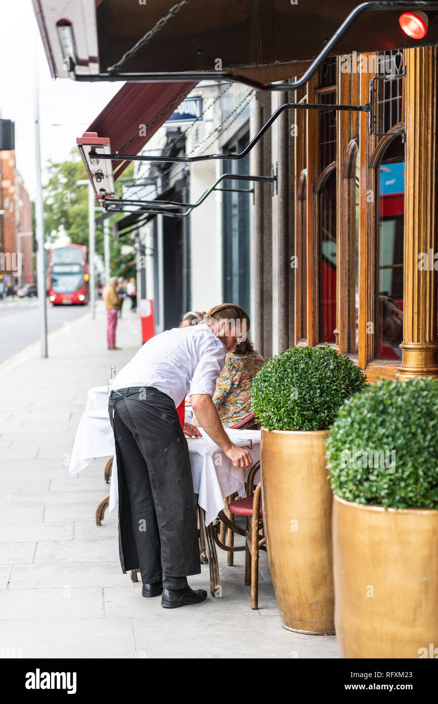 London, UK - September 16, 2018: Man waiter standing setting up cafe dining table in Chelsea outside restaurant street sidewalk during day Stock Photo