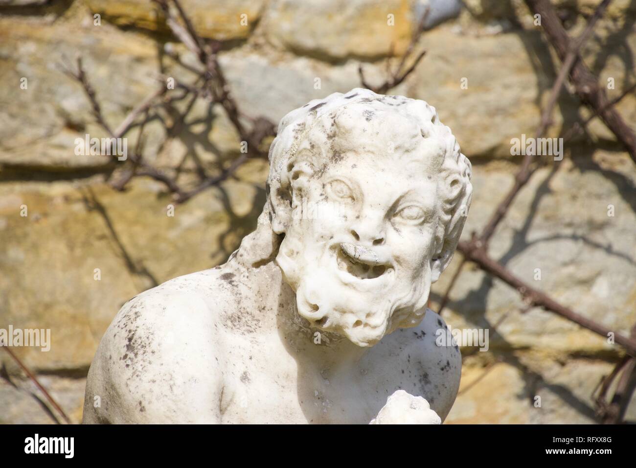 A stone statute, outdoors and in bright sunlight, of a funny old man with a shrivelled face and a big laughing grin that makes him look like a satyr Stock Photo