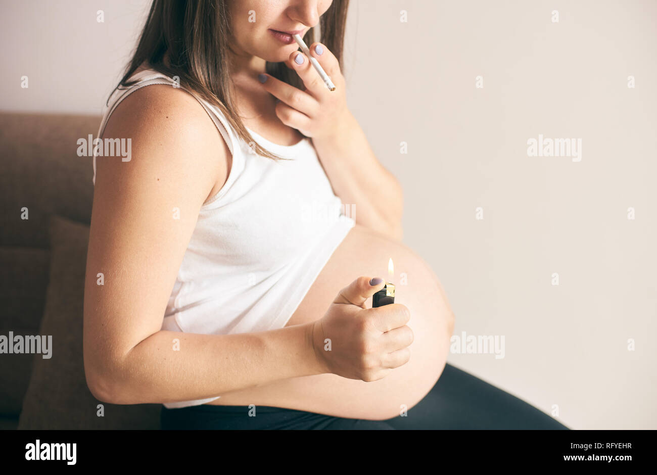 Cropped view of lighter and cigarette in hands of pregnant woman. Female in white shirt smoking during pregnancy period. Concept of threat and harmful Stock Photo