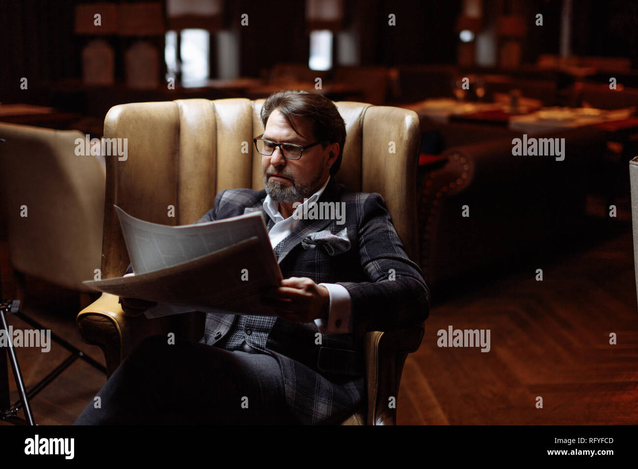 Mid-aged well-dressed man sitting in arm-chair in living room reads newspaper Stock Photo