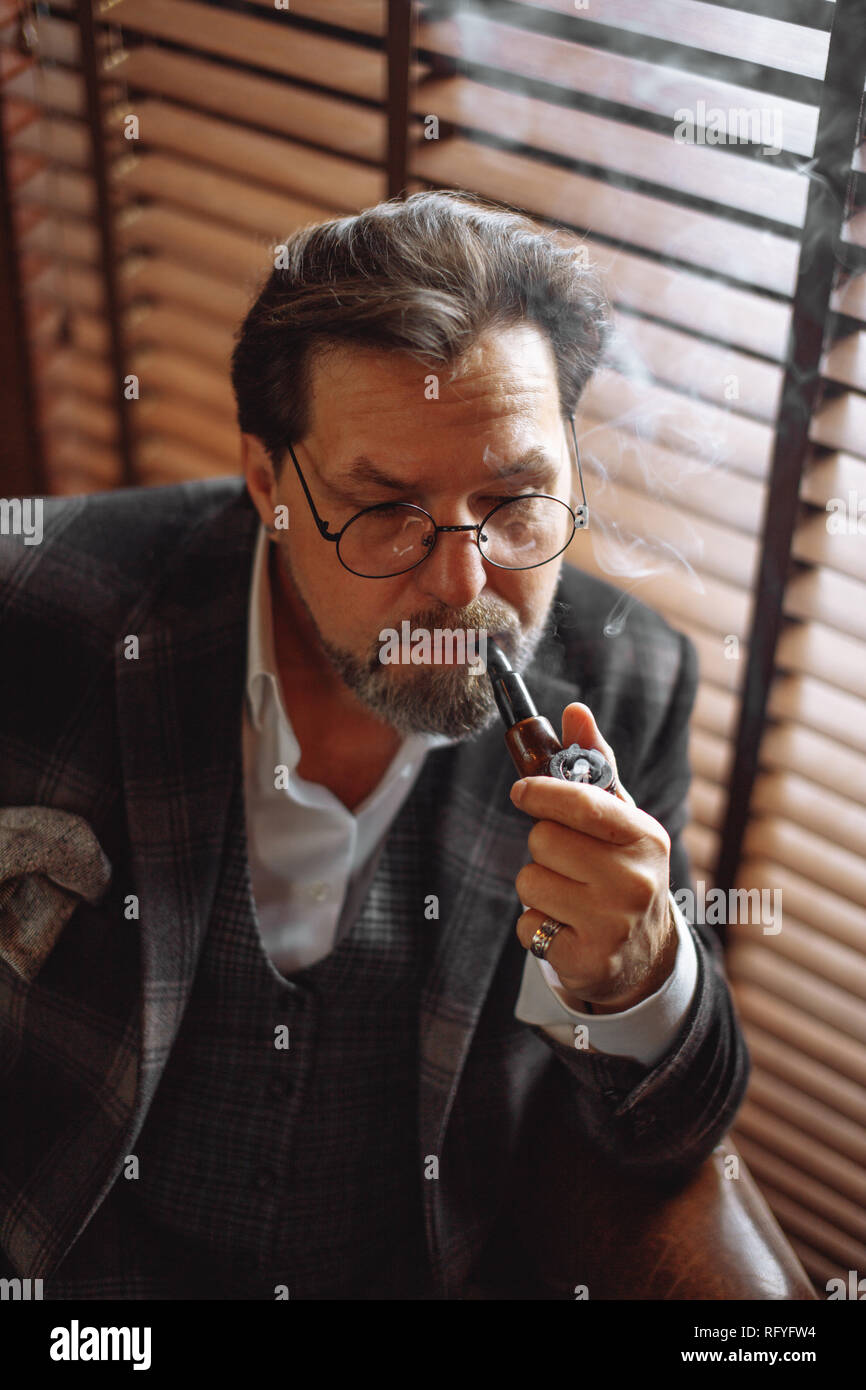Reflective well-dressed mature bearded man in glasses smoking tobacco pipe  Stock Photo