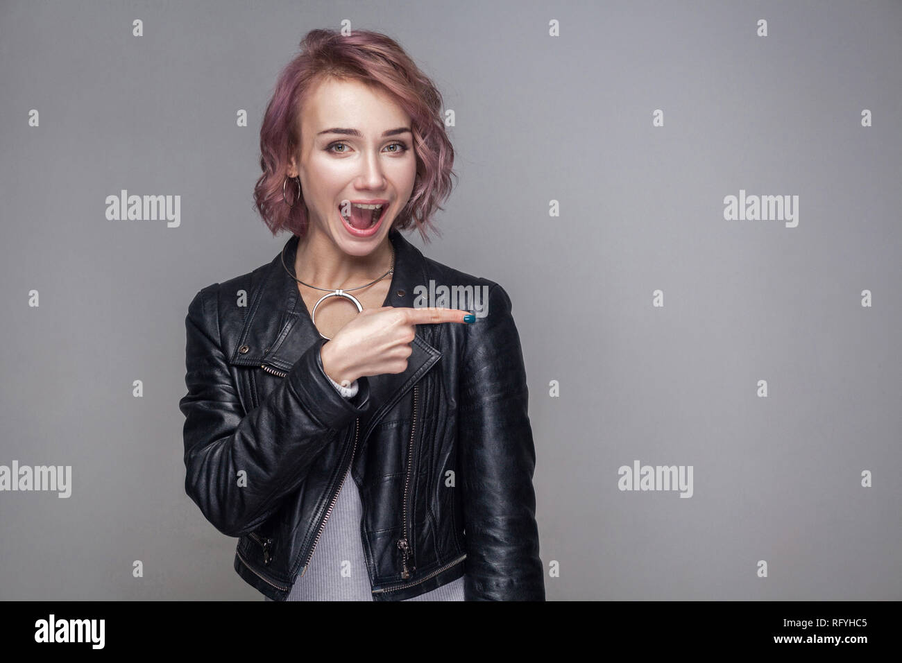 Portrait of surprised beautiful girl with short hairstyle and makeup in casual style black leather jacket standing, looking and pointing at copyspace. Stock Photo