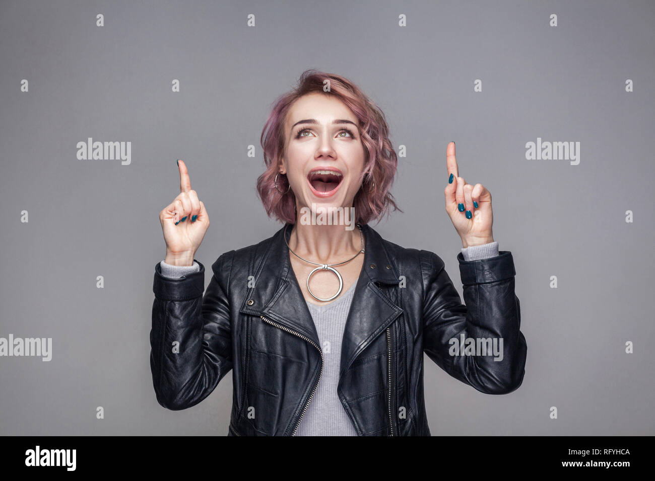Portrait of amazed beautiful girl with short hairstyle and makeup in casual style black leather jacket standing, looking up and pointing at copyspace. Stock Photo