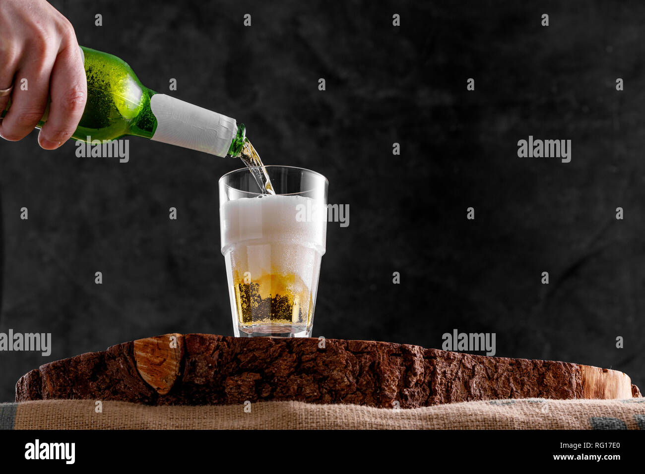 A hand is pouring fresh beer from a bottle into a glass goblet on a wooden background. Close-up. Copy space. Stock Photo