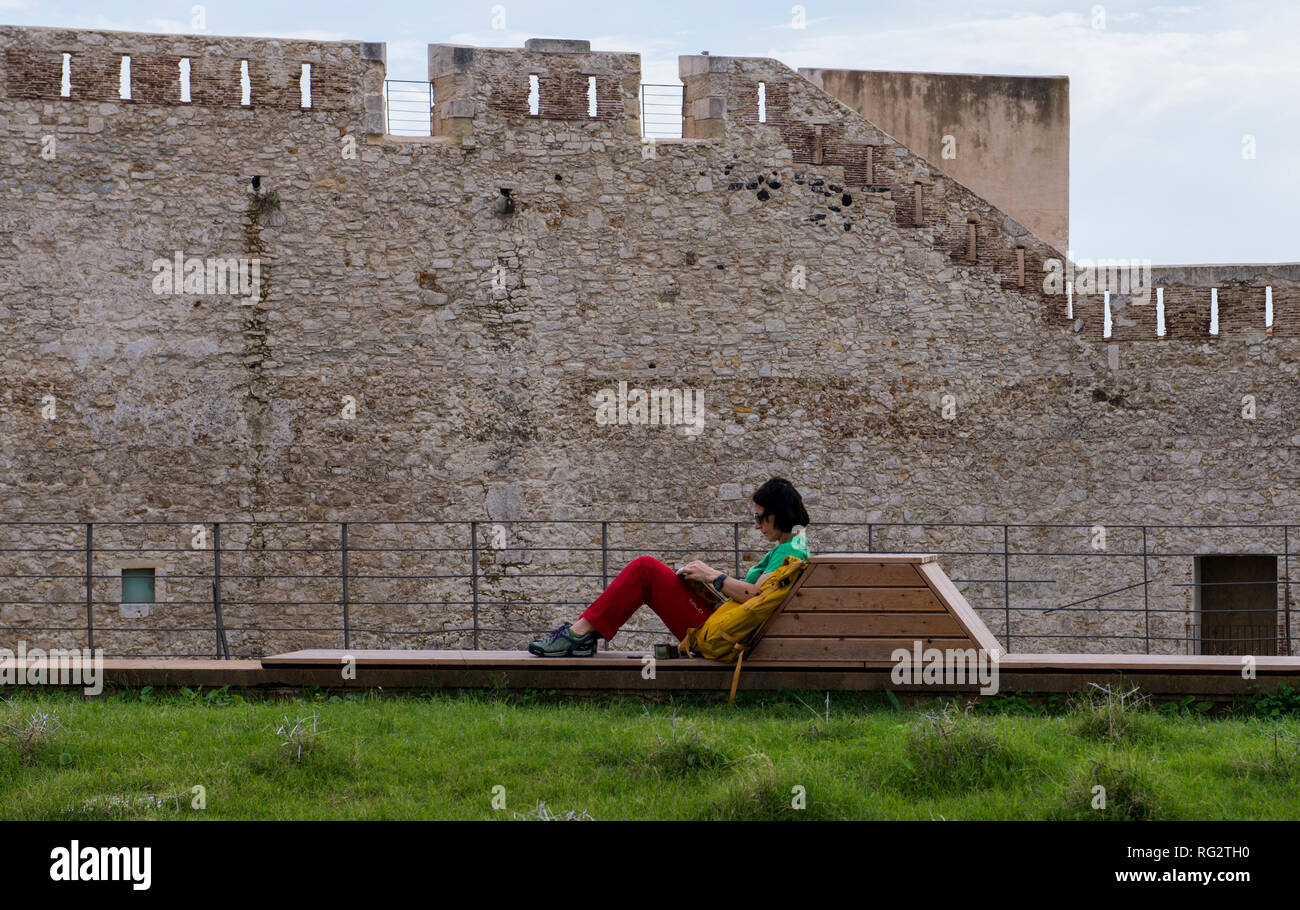 Woman relaxing on wooden bench, fortress wall of The Castello Maniace in background, Syracuse, Sicily, Italy, Europe Stock Photo