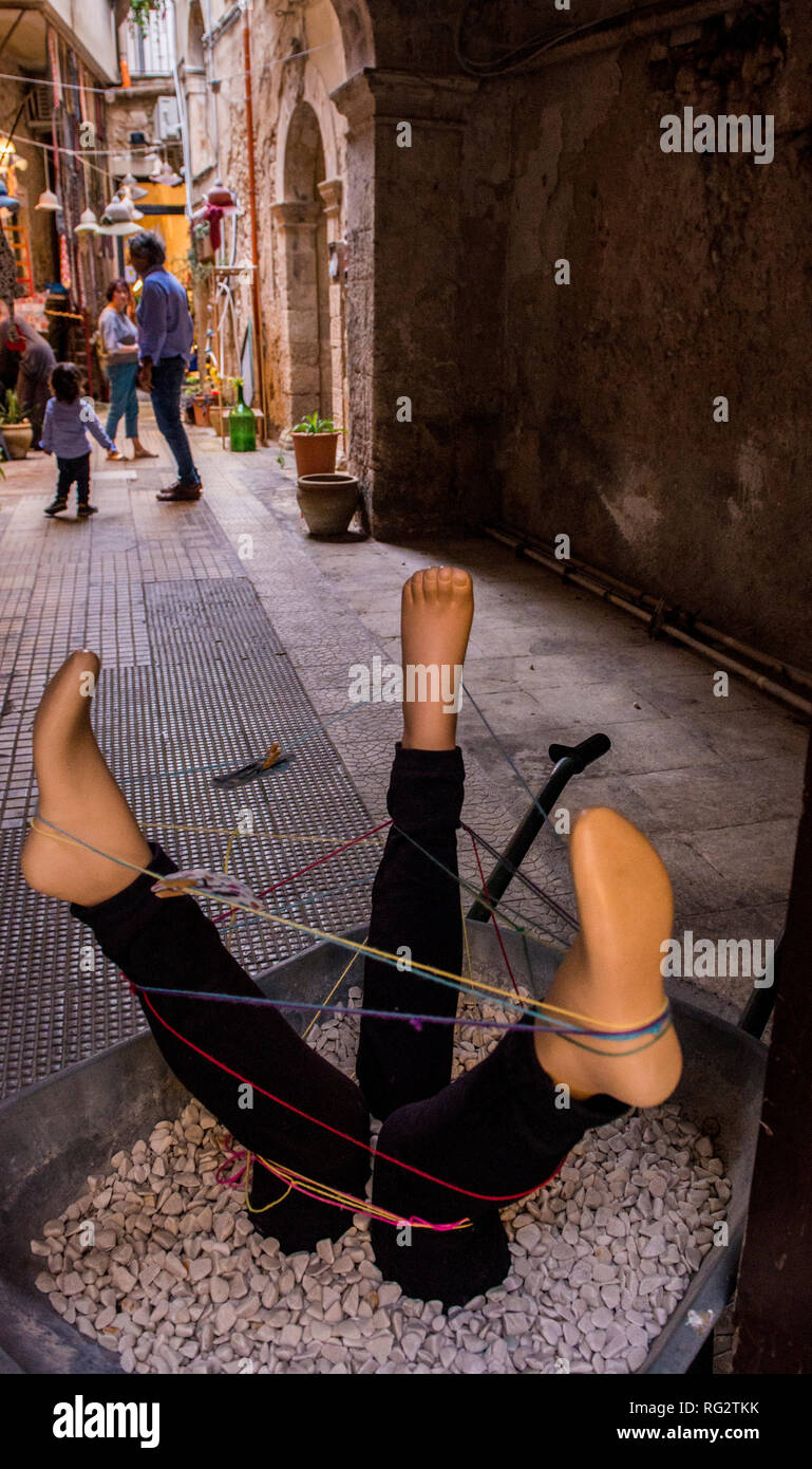 People walking down narrow street, artistic sculpture made of three dummy legs and string in foreground, Ortygia, Syracuse, Sicily, Italy, Europe Stock Photo