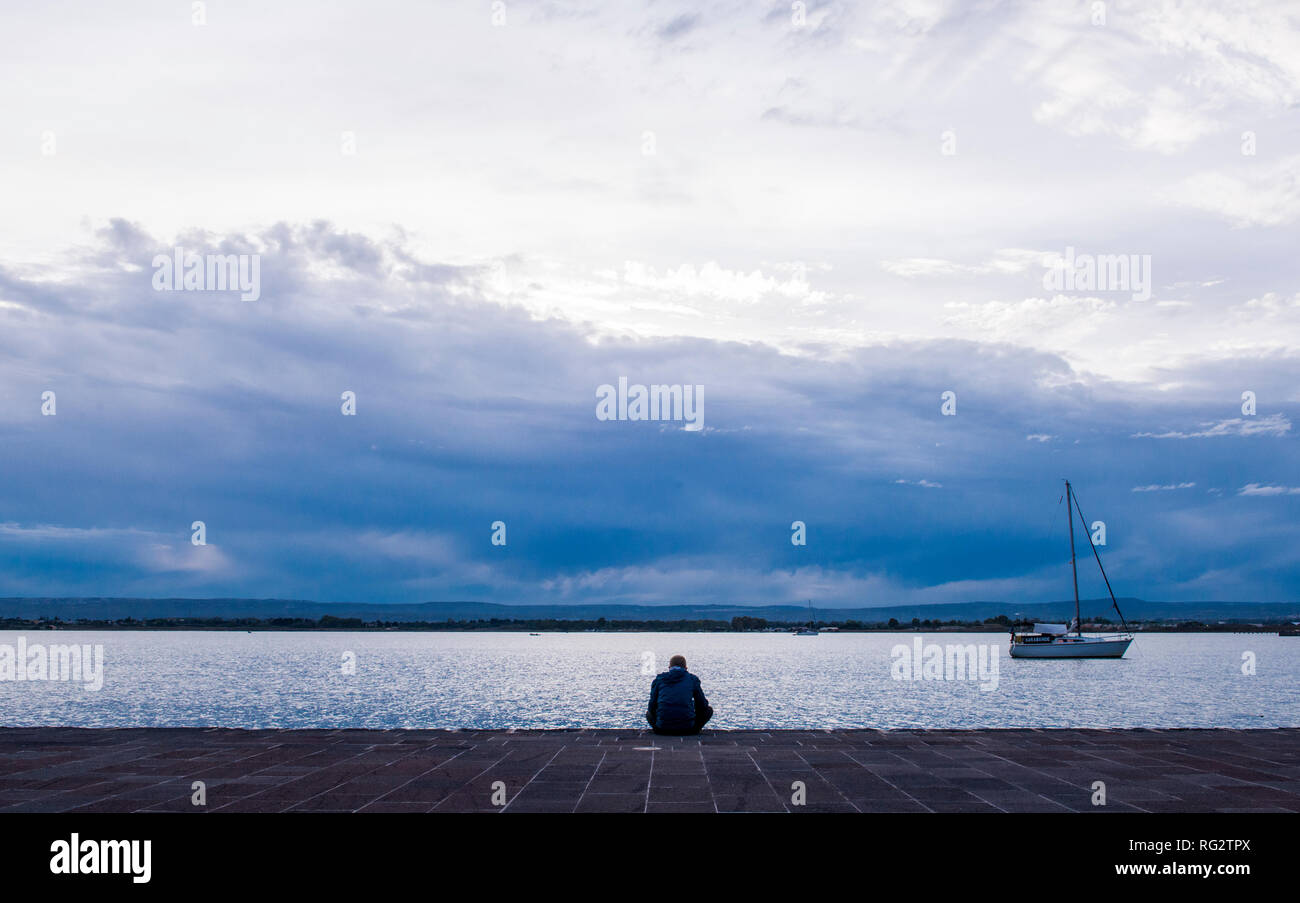 Man sitting beside sea, single boat on water, rear view, Ortygia, Syracuse, Sicily, Italy, Europe Stock Photo