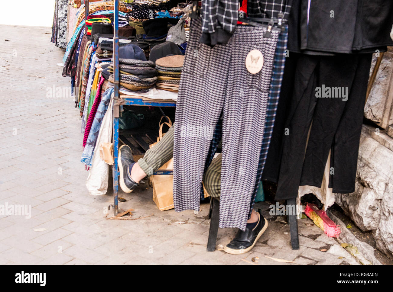 Man sitting beside market stall, feet poking out from behind hanging trousers, Palermo City, Sicily, Italy, Europe Stock Photo