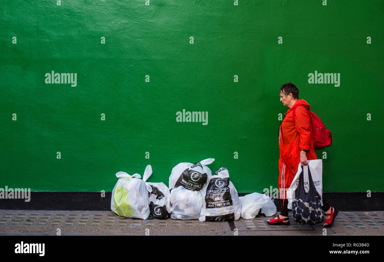Woman in orange raincoat, walking with bags of shopping, past green painted wall, with bags of rubbish in front of wall, Central London, England, UK Stock Photo