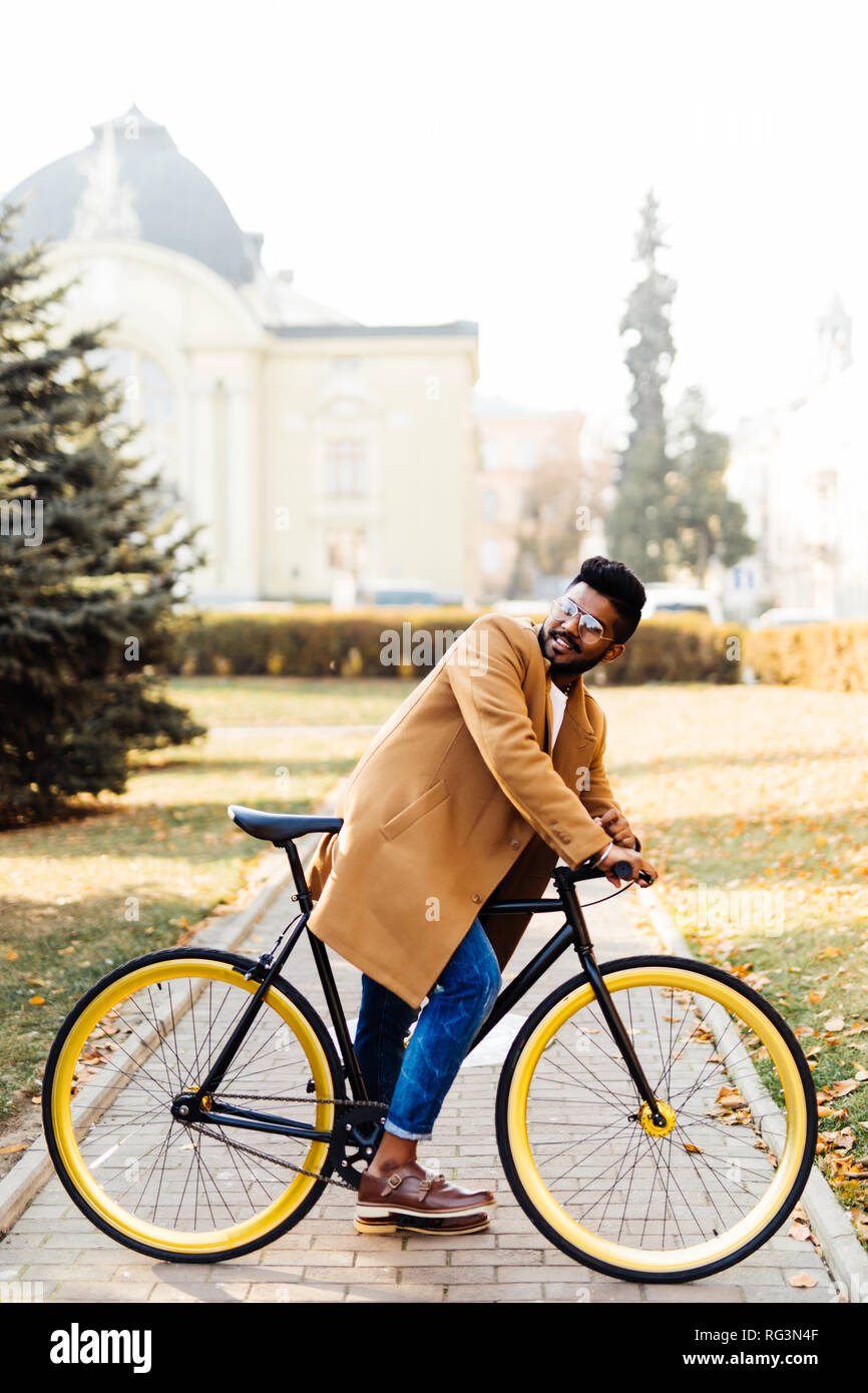 Young bearded indian man in coat while sitting on his bicycle outdoors Stock Photo