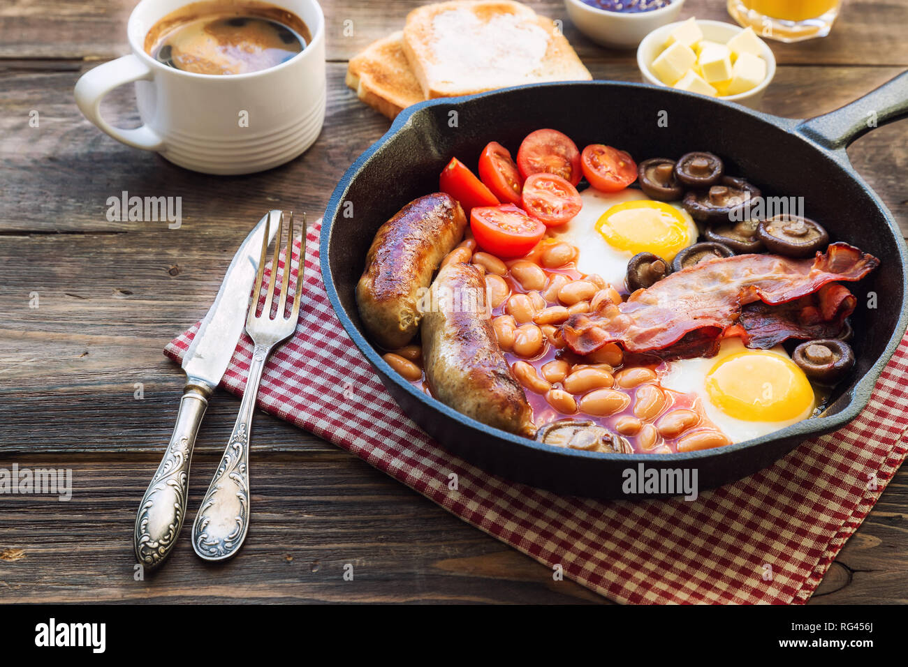 Fried eggs, sausages, bacon, beans and mushrooms in iron skillet, toasts, coffee, butter and jam on rustic wooden background. Full english breakfast.  Stock Photo