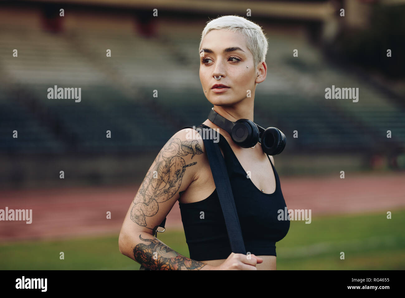 Female athlete standing inside a track and field play ground. Side view of a woman in fitness wear with a wireless headphone on neck looking away. Stock Photo