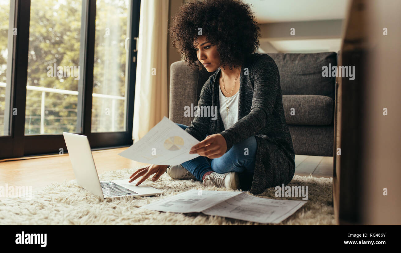 African woman sitting on floor at home with document in hand using laptop. Woman with curly hair working from home. Stock Photo