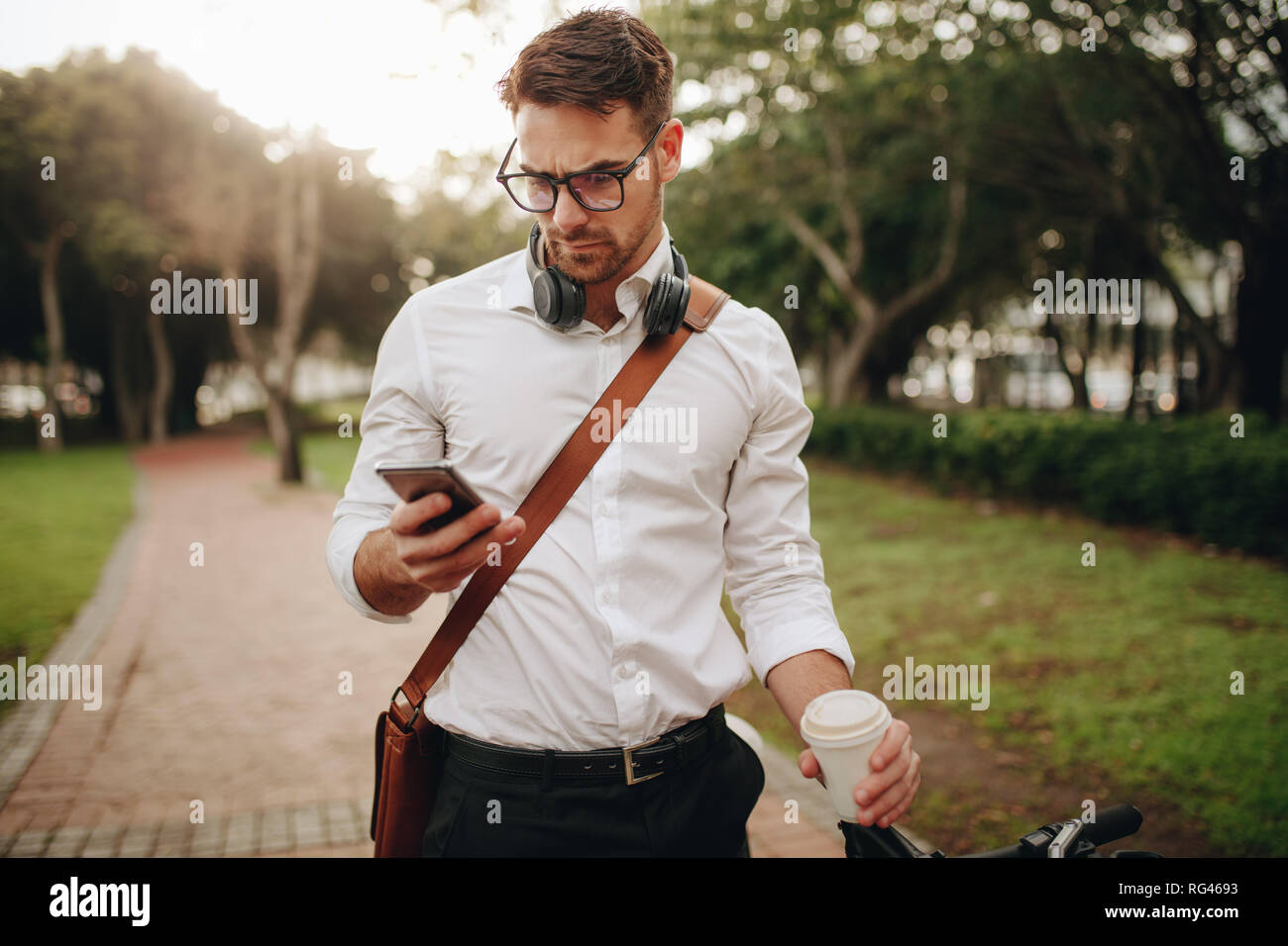 Man standing in park looking at his mobile phone holding a disposable coffee cup. Man walking on street to office carrying office bag and headphones h Stock Photo