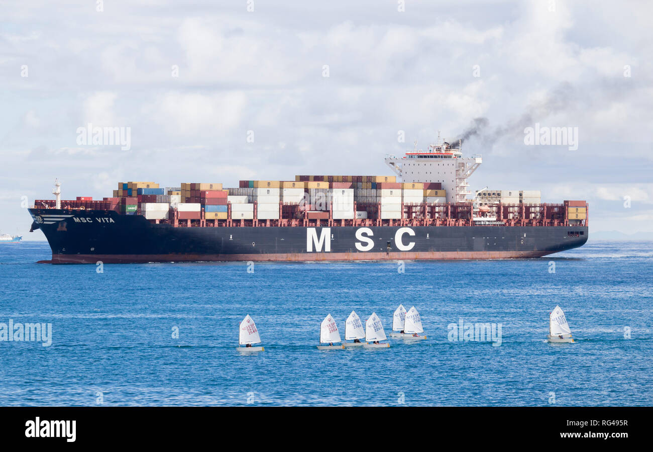 Container ship emitting black smoke passing small sailing boats as it enters port. Stock Photo