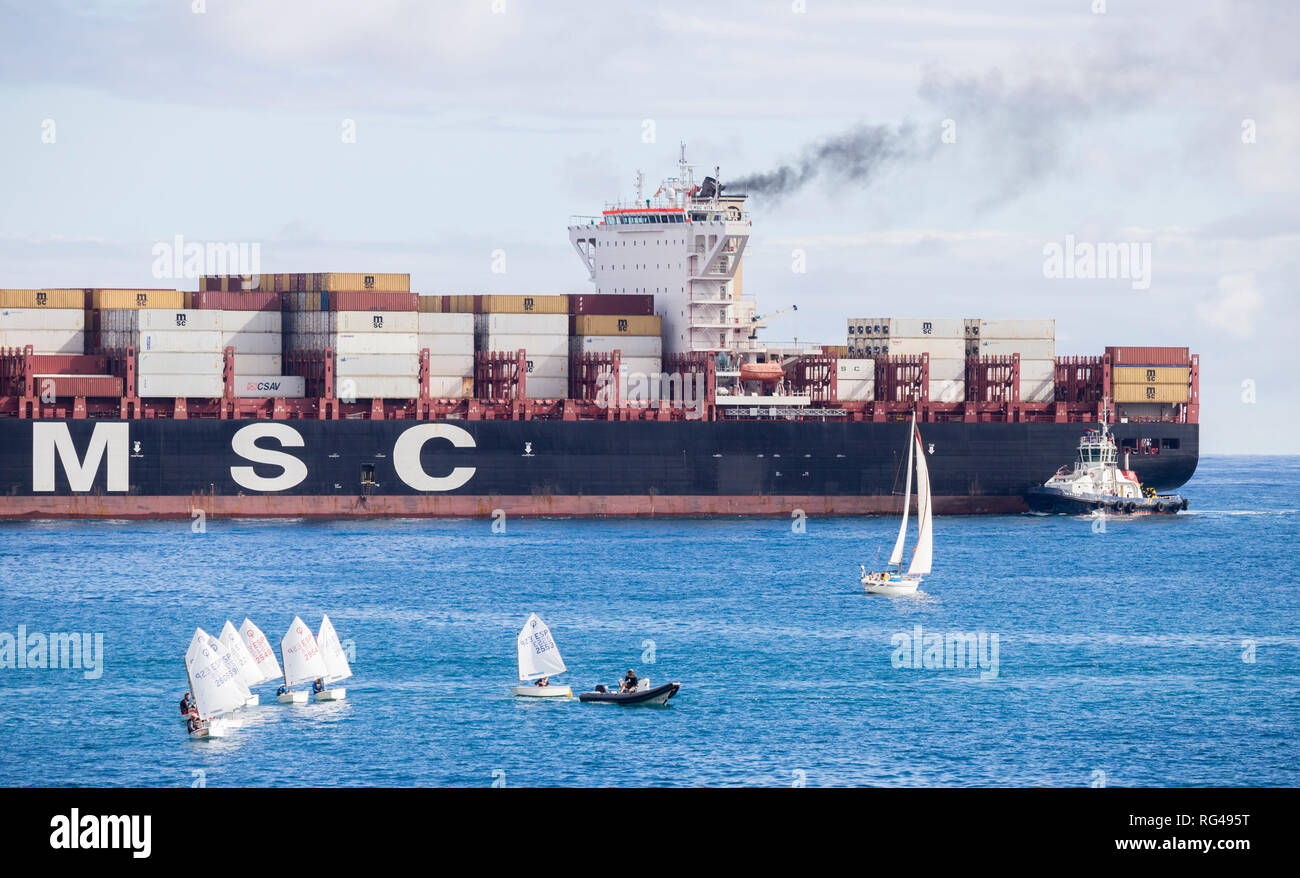 Container ship emitting black smoke passing small sailing boats as it enters port. Stock Photo