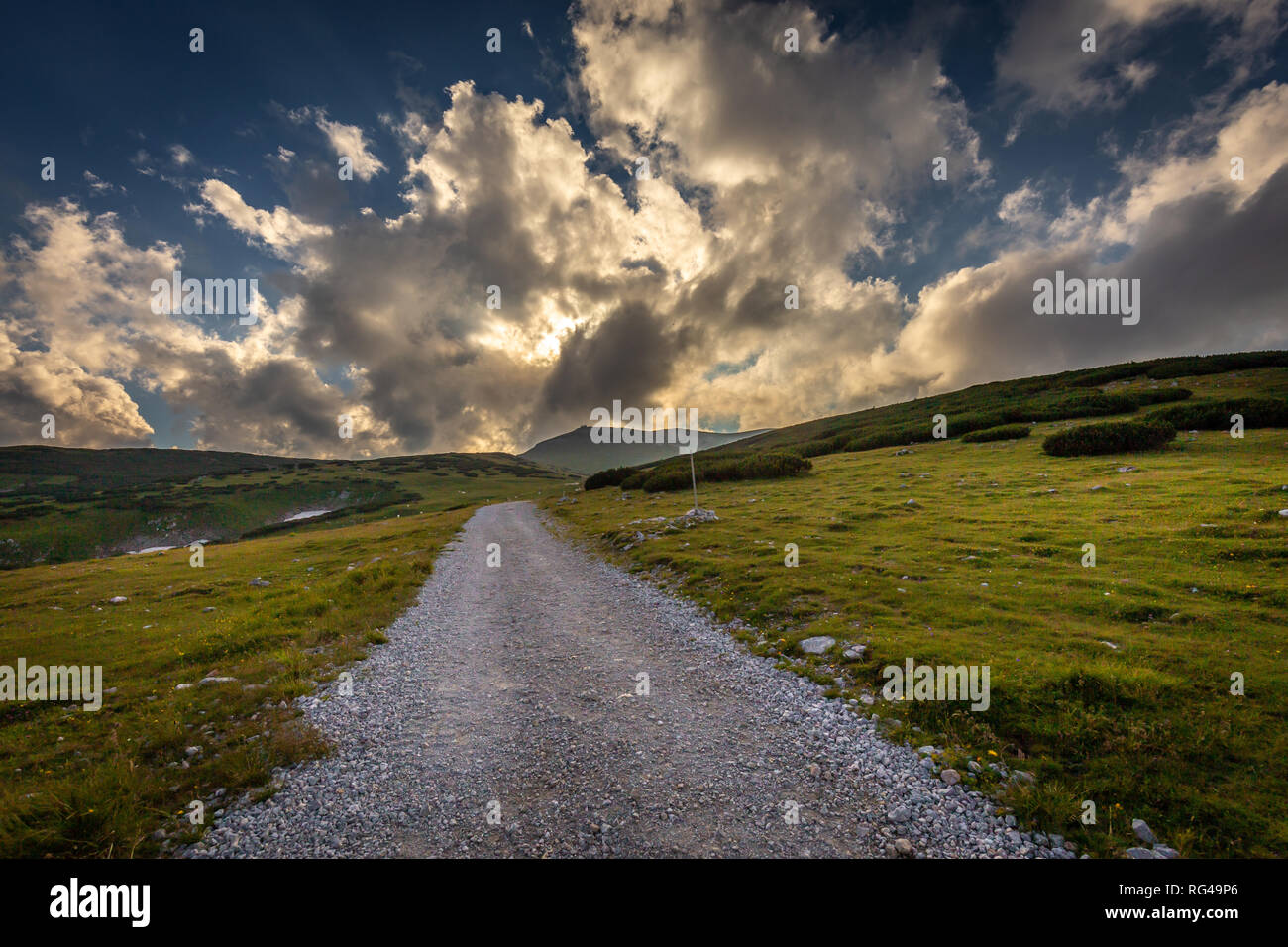 Dirty gravel road through fresh, green, grassy Alpine meadow to Dambockhaus and Fischerhutte from Puchberg, with scenic cloudy dark blue sunset sky Stock Photo