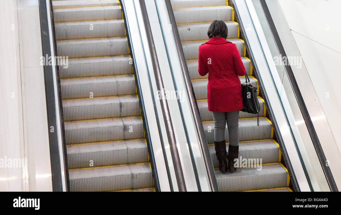 A brunette with a bright red coat riding an escalator up to a second floor with her back to the camera. Stock Photo