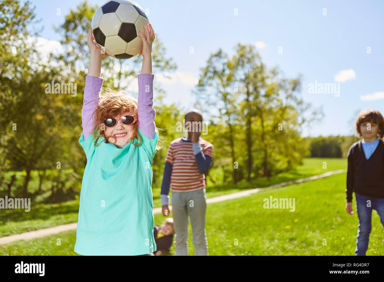 Girl plays football with friends on a meadow in the summer and proudly holds the ball Stock Photo