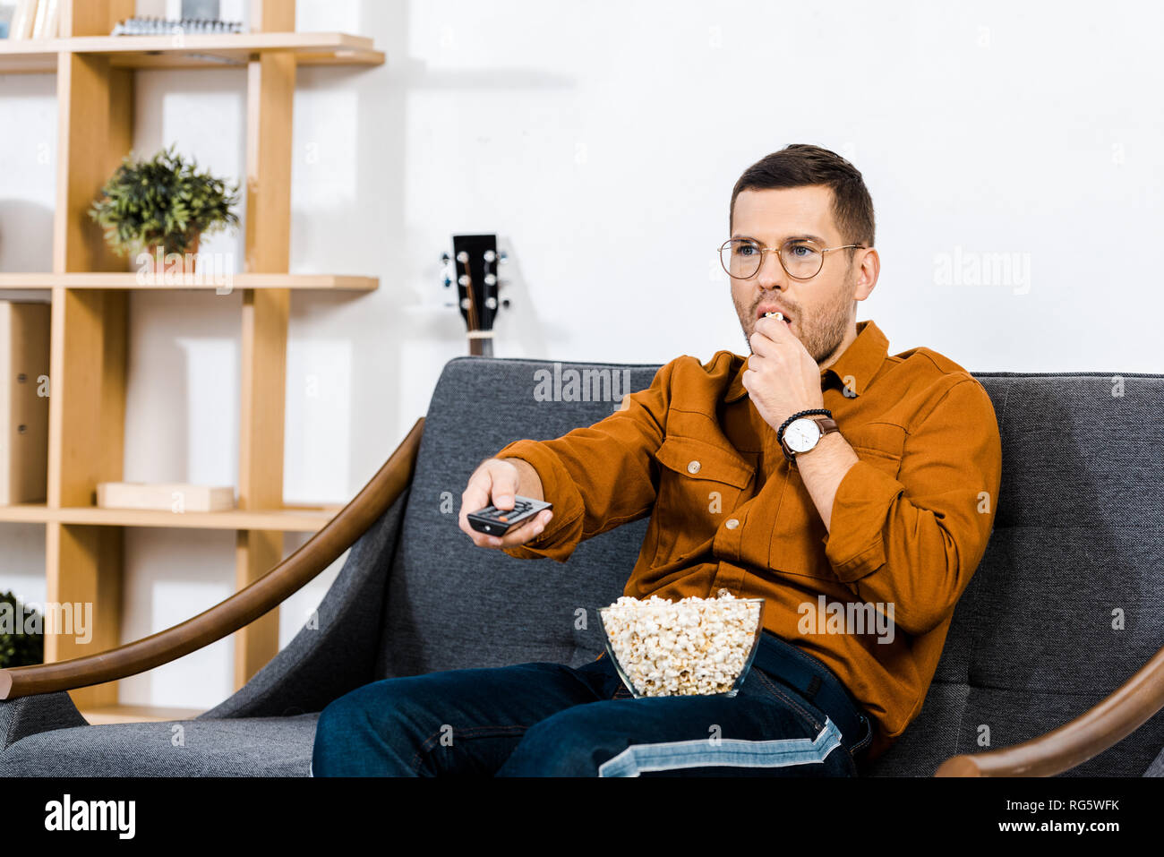 handsome man sitting on sofa, holding remote control and eating popcorn ...