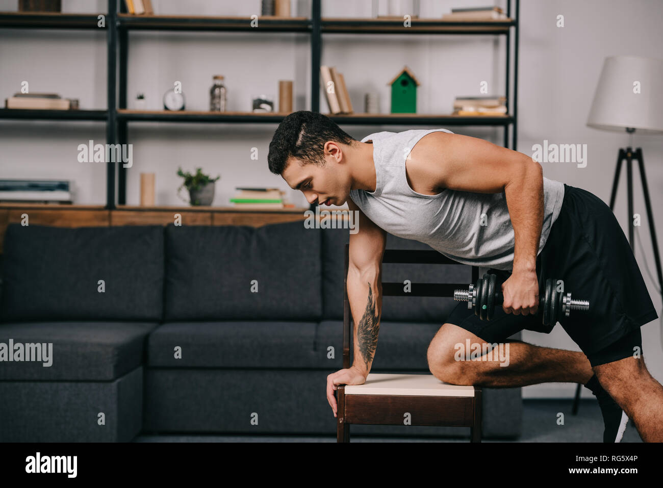 tattooed bi racial man doing exercise with dumbbell on chair in living room Stock Photo Alamy