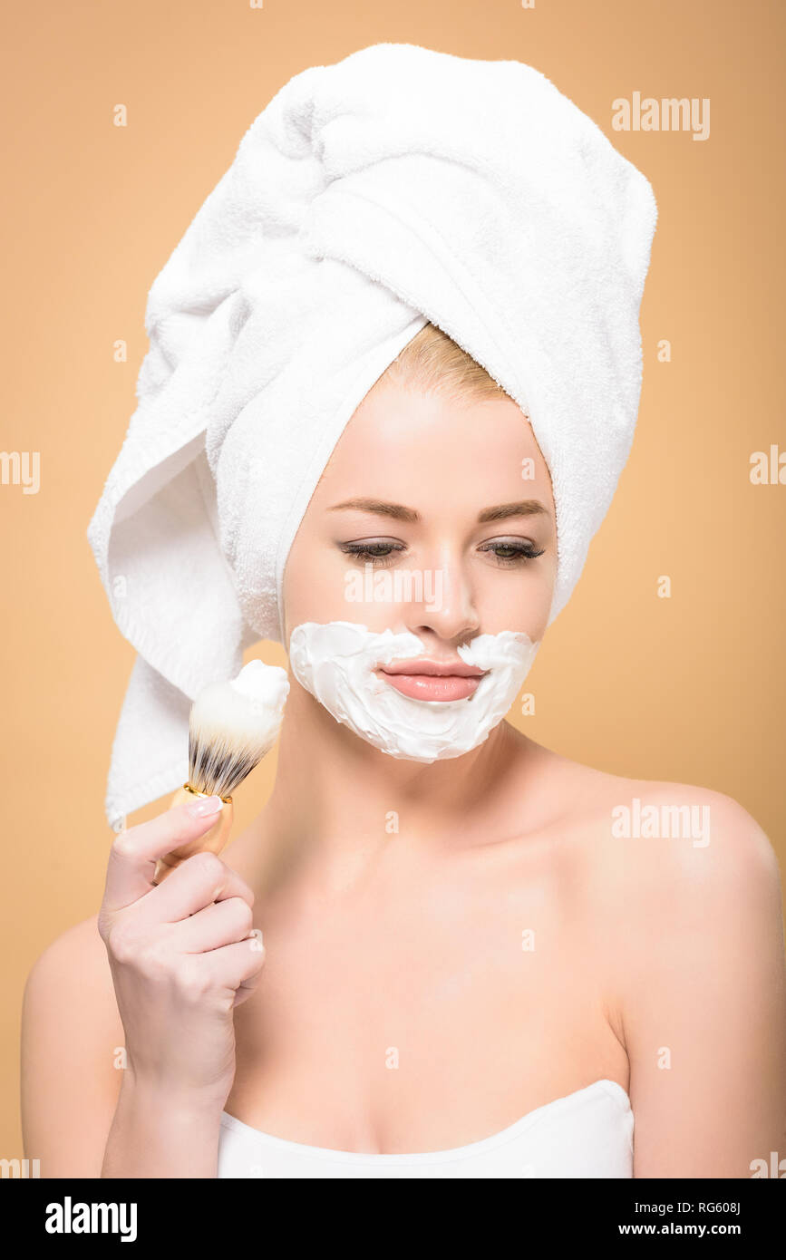 woman with towel on head and shaving cream on face holding shaving brush and looking down