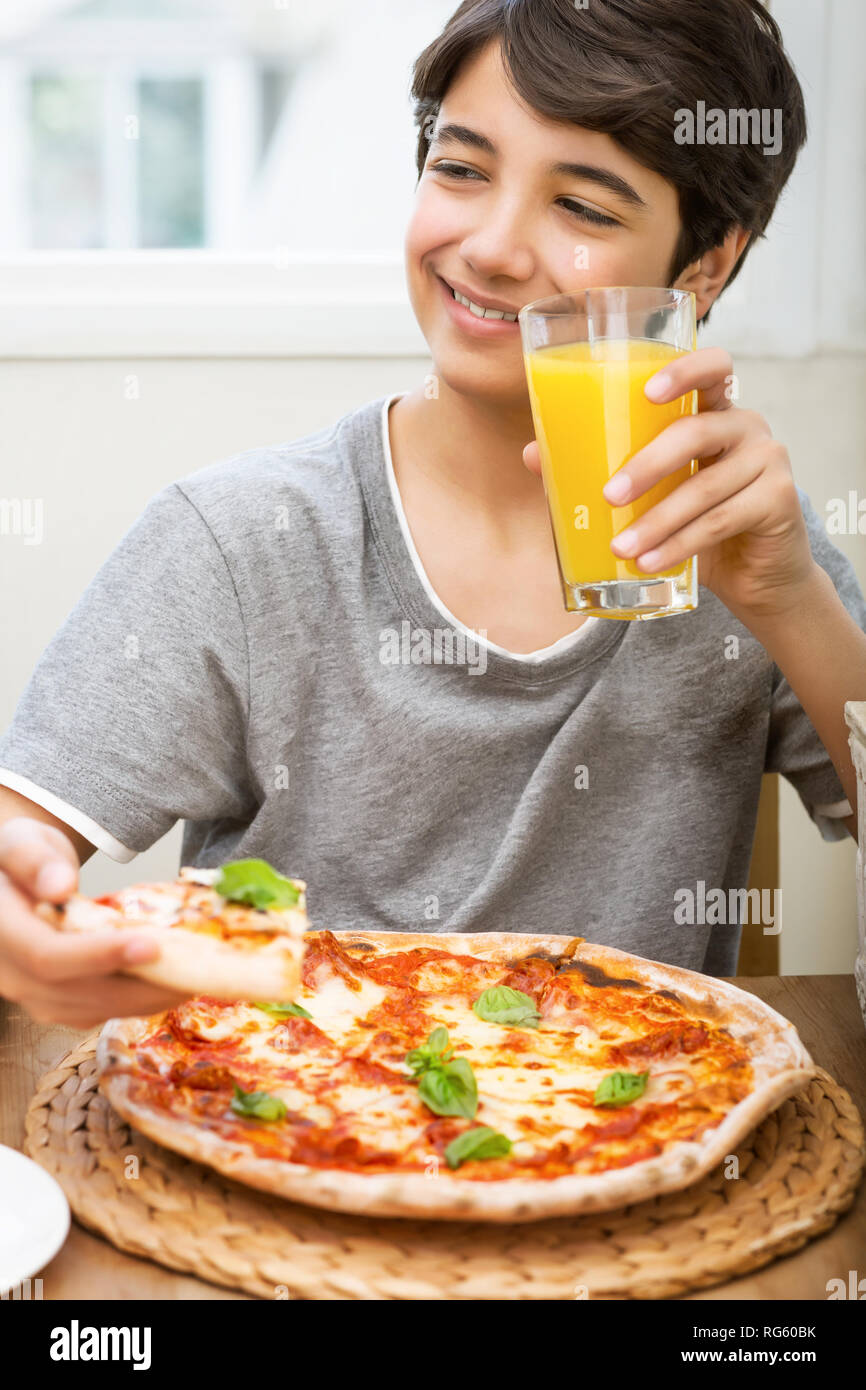 Portrait of a happy teen boy with pleasure eating pizza and drinking orange juice, cheerful adolescent spending leisure time at pizzeria, tasty but un Stock Photo